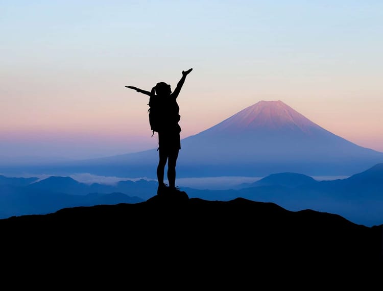 A female hiker is standing on a mountaintop at dusk with her hands raised to the sky.
