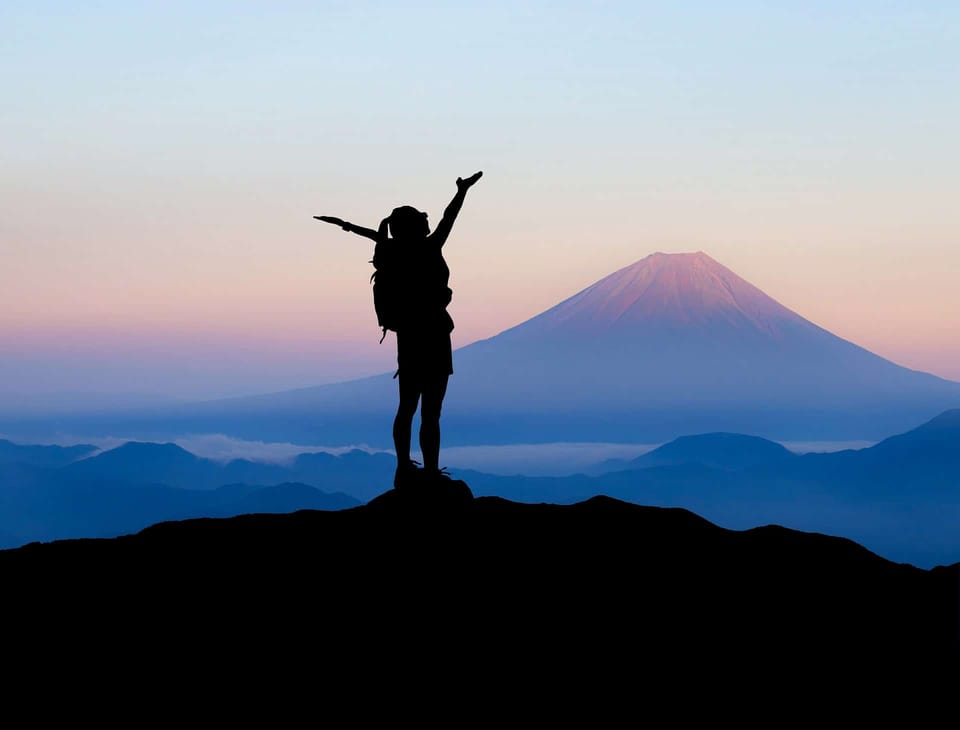 A female hiker is standing on a mountaintop at dusk with her hands raised to the sky.